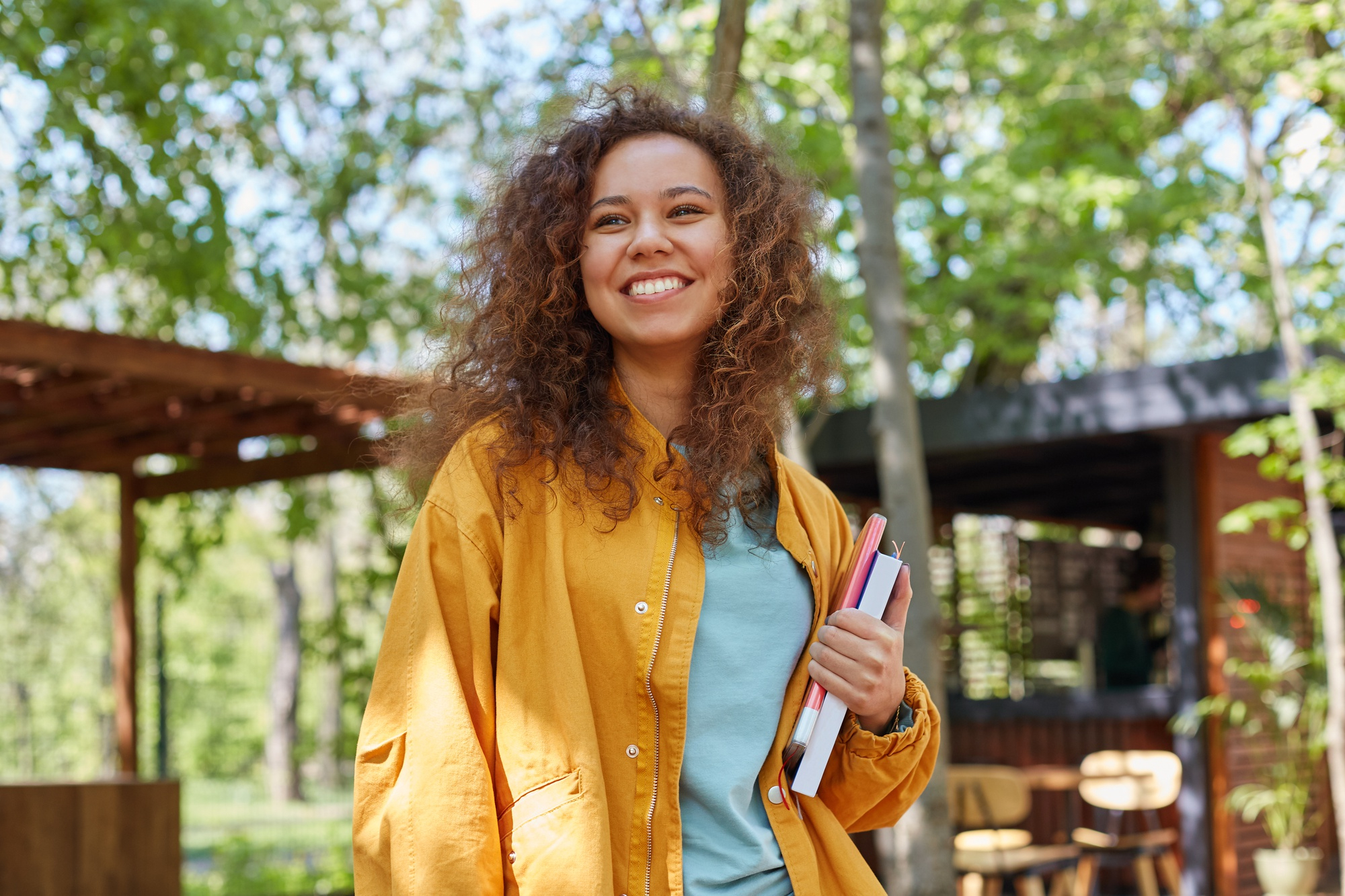 portrait-smiling-young-beautiful-dark-skinned-curly-student-girl-cafe-terrace-holding-textbooks-wearing-yellow-coat-enjoys-weather